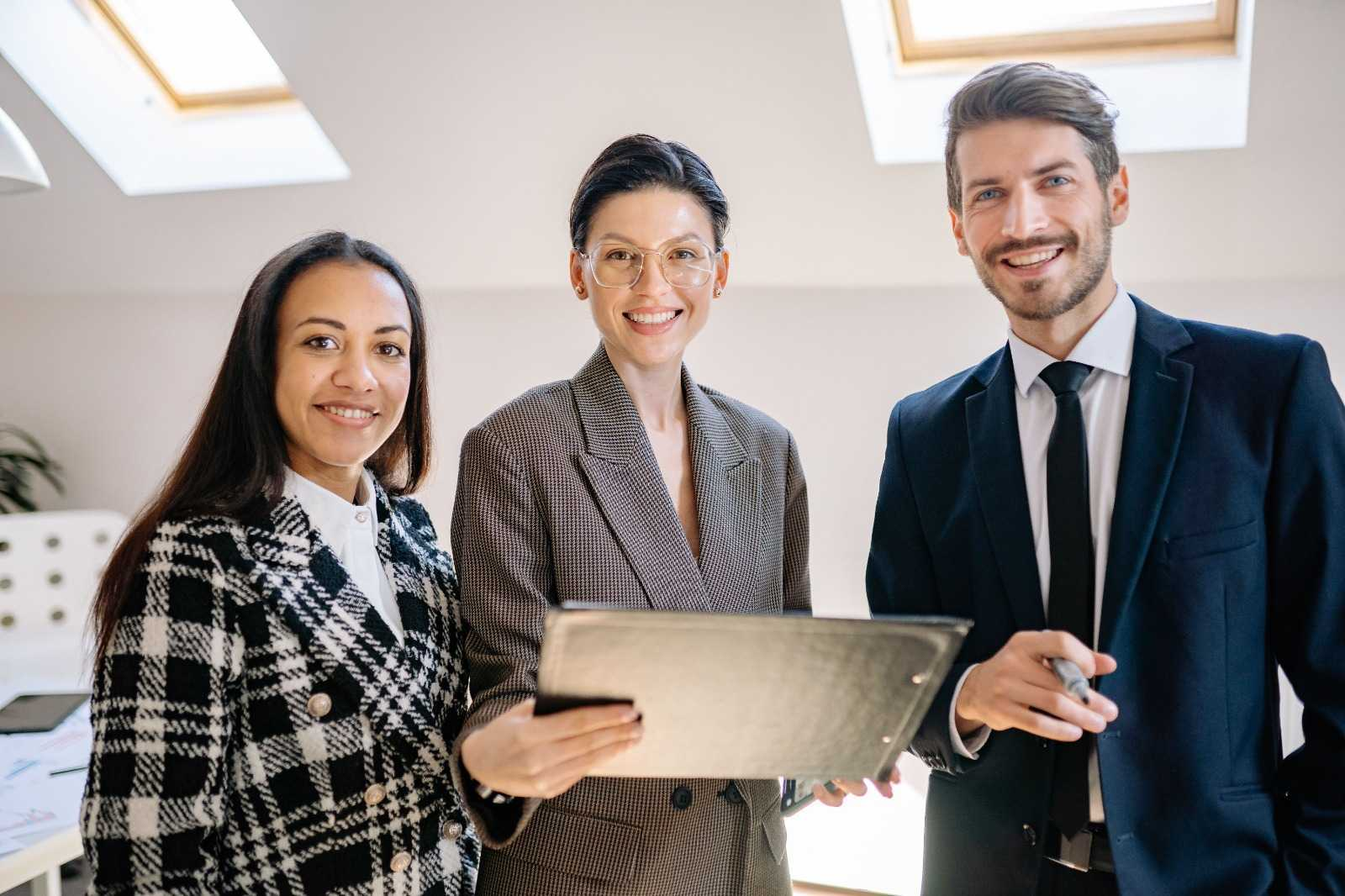 Three smiling professionals standing together with a document folder in a bright office.